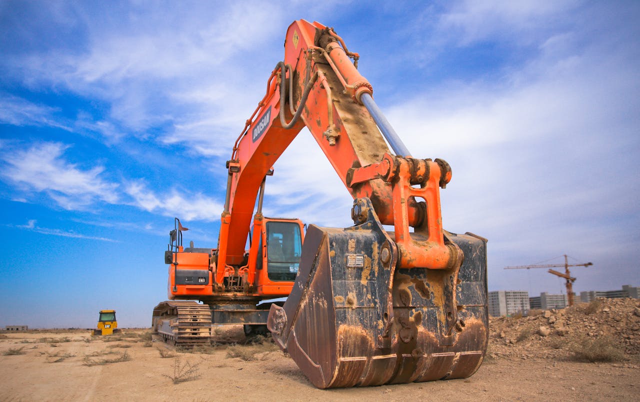 impact-img A large orange excavator working on a construction site under a blue sky.